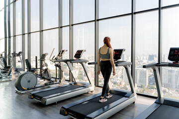 Woman walking on treadmill in modern high-rise gym with large glass windows and panoramic urban skyline, focusing on wellness and fitness lifestyle