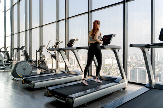 Woman using treadmill in modern high-rise gym with panoramic city view, engaging in indoor cardio workout and healthy lifestyle fitness training routine
