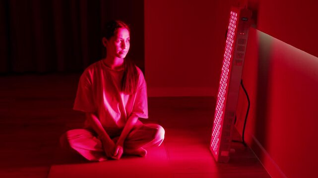 Young white woman sits cross-legged with closed eyes, relaxing in front of a red light therapy panel in a calm home environment