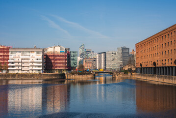 Fototapeta premium The image shows Liverpool's Albert Dock with red brick architecture, modern glass buildings in the background, calm water, and a small central bridge.