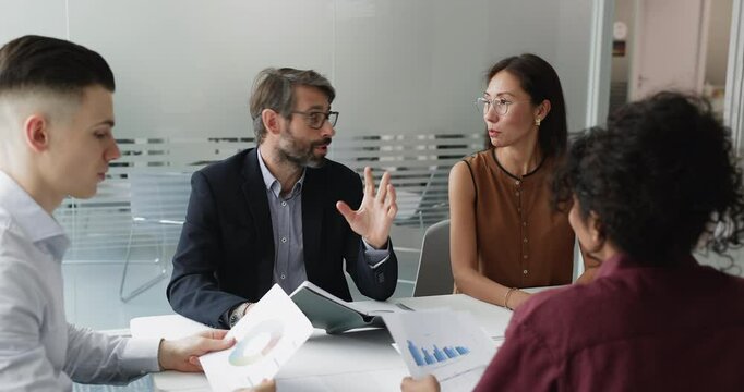 Middle-aged team leader shares analytical information to diverse young staff members seated at table in modern meeting room, discussing project progress or performance metrics, considering data trends - Powered by Adobe