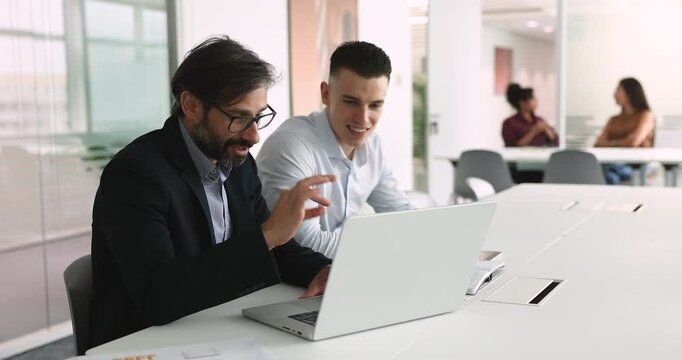 Two male colleagues focused on laptop screen, older man guiding or explain new software to younger man listens attentively engaged in discussion, review data, analyze report, finalizing project detail
