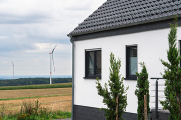 Modern single-family homes near the edge of open fields. Wind turbines rise in the background, symbolizing the contrast between rural living and energy transition.