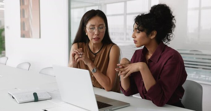 Two multiethnic women teammates working on joint online tasks seated at table in modern office, analyzing results, review project presentation, discussing updates or new AI business platform on laptop