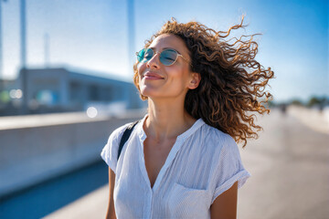Woman with curly hair blowing in wind, smiling with sunglasses in bright sunlight. Wearing blue striped shirt in urban setting. Expression of happiness, joy, contentment and carefree freedom. Natural