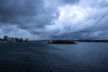 Image of small island in the middle of Halifax Nova Scotia Canada harbor with small Light house lit up cityscape and dramatic cloudscape.