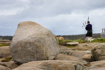 Photo of are large granite boulder and back of a man playing bagpipes Location Peggy's Cove Nova...
