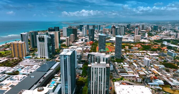Flight over the tops of the high-rise buildings in the cityscape of Waikiki, Honolulu, Hawaii, USA. Azure waterscape and cloudy sky at backdrop. Aerial view.