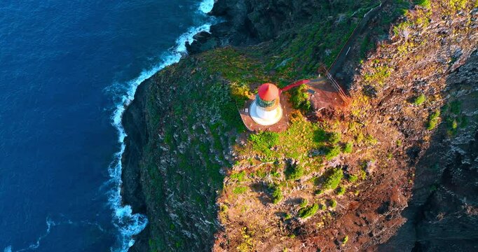 Footage above the red roof of the light house standing on the top of the steep rock. Waves splash by the rocky shore. Aerial view.