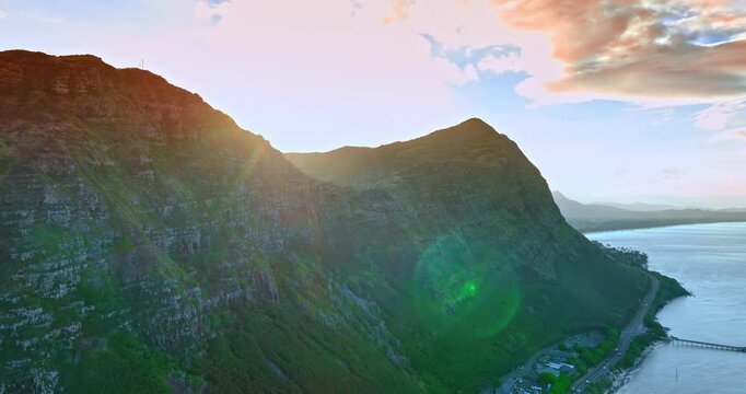 Bright sun dazzling the view of the Huge massive bare rock with highway at the foot. Drone footage above the inhabited area at the rocky shore. Hawaii, USA.