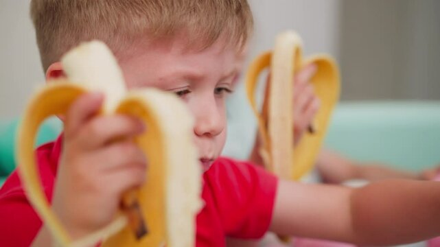 Close up of young boy eating banana while sitting in pastel ball pit, playing and enjoying snack time with blurred view of another child also eating banana and playing in cheerful indoor environment