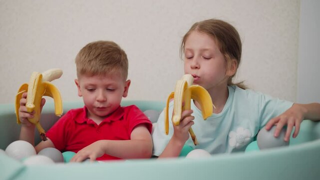 Cute kids sitting together in pastel ball pit eating bananas happily, boy in red shirt holding banana while girl in blue shirt bites into hers, playful mood captured with bright soft light