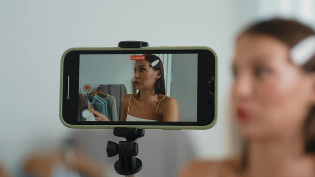 Young social medial content creator woman make fashion video. Blogger smiles to camera using selfie stick and light ring while making persuasive online clothing sell vlog to audience or follower. Adit