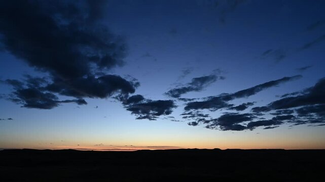 Time lapse of late evening clouds in a blue sky with orange shades above rolling hills in the distance
