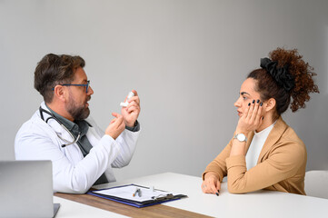 Male doctor in lab coat and glasses explaining how to use inhaler pump to female patient at desk in...