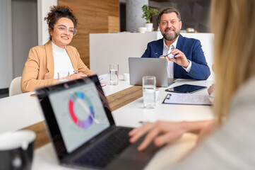 Smiling businessman and businesswoman looking at female colleague presenting report over laptop in meeting