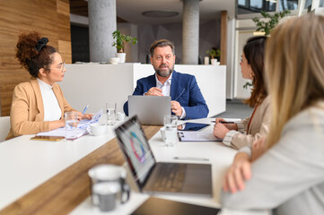 Obraz premium Male manager with laptop on table presenting data and reports to female professionals in meeting at board room