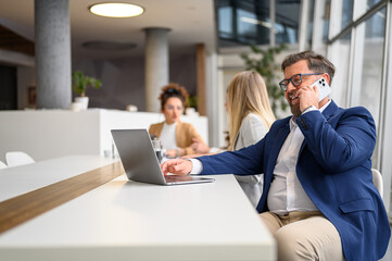 Businessman working online on laptop while discussing over smart phone at desk with colleagues in the background