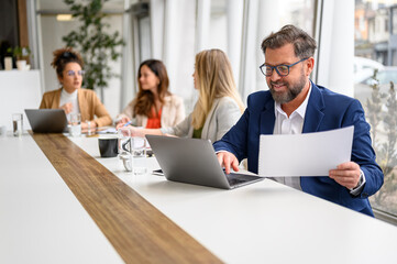 Fototapeta premium Male accountant reviewing financial documents and using laptop with female professionals in the background