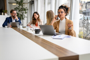 Female accountant discussing on mobile phone and working over laptop while sitting with coworkers in the board room