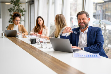 Businessman holding glasses and analyzing reports over laptop while sitting by female professionals in board room