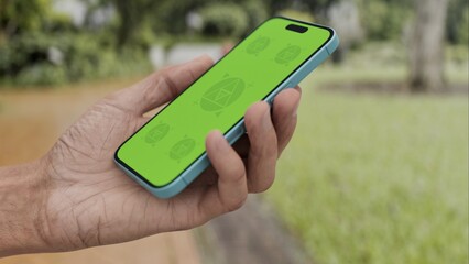 GREEN SCREEN CHROMA KEY Person holding a smartphone with a green screen in a lush garden environment, surrounded by greenery and a scenic path during daylight