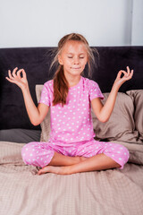 Little beautiful girl meditating in lotus position sitting on bed in cozy bedroom. Healthy lifestyle. Hobbies. Wellness.
