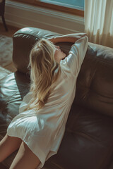Beautiful blonde woman in elegant white robes reclining on a leather couch in cozy sunlight.
