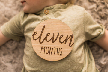 adorable 11 month old baby boy laying with a milestone wooden round sign on him