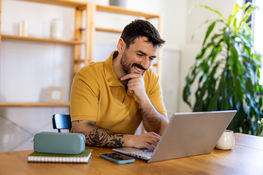 Young man working from home office using laptop and smiling, enjoying remote work and flexible hours