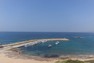 Fishing boats are anchored in a serene harbor surrounded by clear blue waters. The warm sun shines on the coastline, revealing rocky formations and a tranquil atmosphere.
