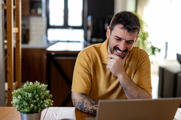 Happy male freelancer laughing while working on a laptop in a cozy home office, enjoying the...