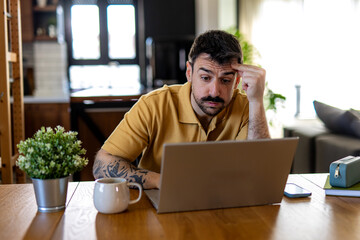 Young man looking worried while working at home with his laptop, having a cup of coffee next to him