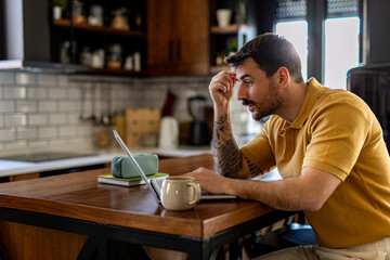 Freelancer working from a cozy home kitchen, focused on his laptop while expressing concern over ongoing work challenges