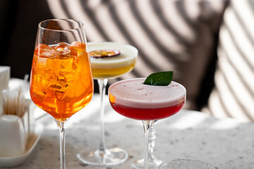 Three beautifully crafted cocktails are displayed on a marble surface, featuring vibrant colors. The drinks include an orange spritz, a creamy yellow blend, and a red beverage garnished with a leaf.