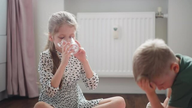 Little girl with long blond hair in spotted dress drinking water from clear glass cup while little boy in green shirt sits beside thoughtfully bent over