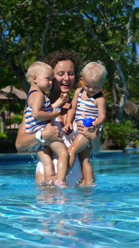 Vertical screen loving mother holding smiling twin toddlers while playing with colorful toys during sunny swimming pool leisure at tropical resort, enjoying family bonding moment
