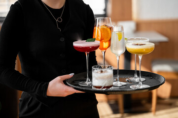 A server presents a tray filled with five vibrant cocktails, each showcasing unique colors and garnishes. The stylish drinks reflect a lively bar atmosphere, perfect for social gatherings
