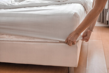 hands of woman making bed, tucking in fitted sheet and bed linen in lighted room with large window....