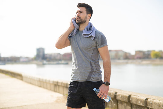 Athletic young man taking a break after workout outdoors with towel and water bottle
