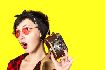 Young sexy brunette woman with bandana and heart shape red glasses with retro vintage film camera against yellow background