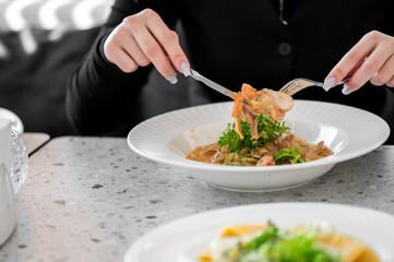 A person uses a fork and knife to enjoy a creamy pasta dish, highlighting delicious ingredients and vibrant greens. The elegant plate is set on a stylish table, perfect for dining.