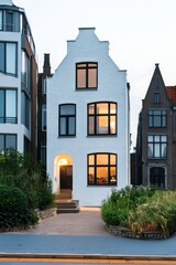 Fototapeta premium Modern house with unique architecture in an urban setting illuminated at dusk with lush greenery and surrounding buildings