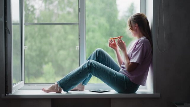 Side view of young woman seated comfortably on window ledge eating pizza slice with plate nearby under soft natural light, peaceful indoor scene with blurred lush green outdoor background of trees