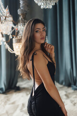 Naklejka premium beautiful brunette with thick hair in a long black dress in a studio with sand and chic chandeliers. Gray walls and floor-to-ceiling windows. Elegant woman of 30 years old, athletic figure. Portrait
