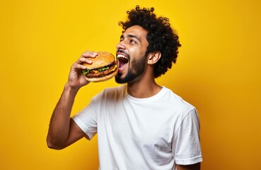 Young Indian man eats large tasty burger with open mouth, isolated on yellow background. Hungry student with curly hair enjoys fast food meal. Cheeseburger snack, lunch, diet, unhealthy eating.