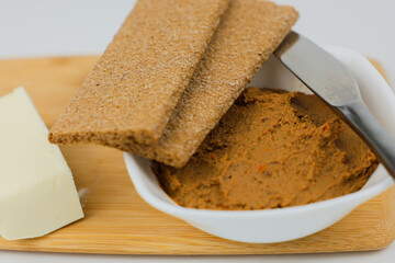 In close-up on a wooden board there is a white plate with liver pate, slices of bread and a knife lie on top and a piece of butter on a white background next to it. 