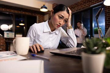 Closeup of professional woman showing signs of exhaustion, sitting at desk, looking at her laptop late at night. Frustrated employee overwhelmed by deadline pressure scrolling through online documents