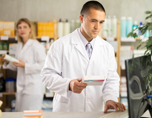 Fototapeta premium Qualified male pharmacist working at a computer against the background of a female pharmacist, they check the availability of medicines in the electronic table and in the pharmacy warehouse