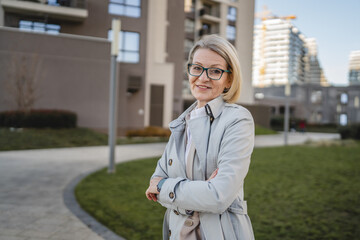 One mature woman stands with folded arms outdoors in front of a building